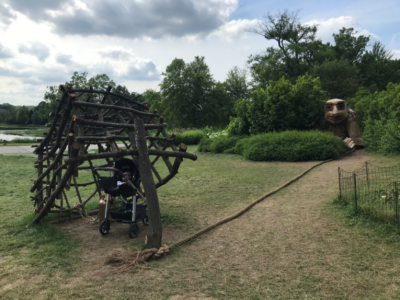 A baby in his stroller sits under a trap made of tree limbs with a giant wooden troll in the background.