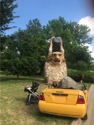 A massive wooden troll holds a boulder over a yellow convertible.