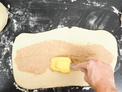 Dough is rolled out thin on a floury surface, with a spatula spreading a filling on it.
