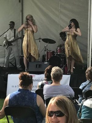 Two women in matching gold dresses sing on a stage with a backup band.
