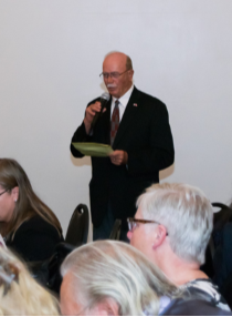 A man in a suit speaks into a microphone in front of seated guests.