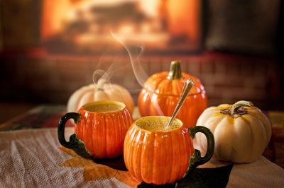 pumpkin-shaped mugs with steam coming from the sit on a table with real pumpkins.