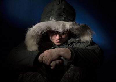 A man wearing a big parka with a fur hood looks down.