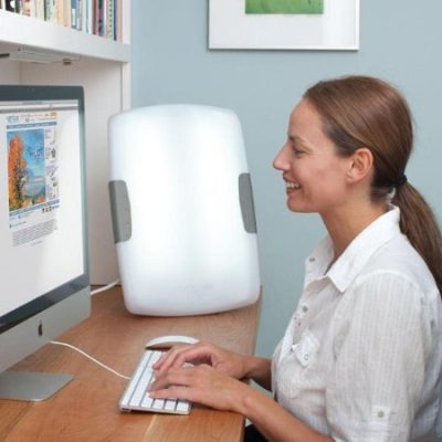 A smiling woman sits at her computer with a light therapy bulb next to her.