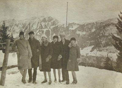 An old sepia photos features men and women in the snow against a snowy mountain range.