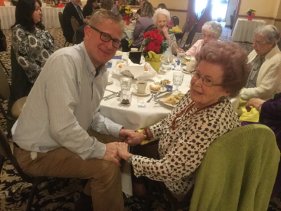 An older couple sit clasping hands at a table in a dining hall.