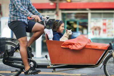 A man drives a bike with a child and dog in a carrier in front.
