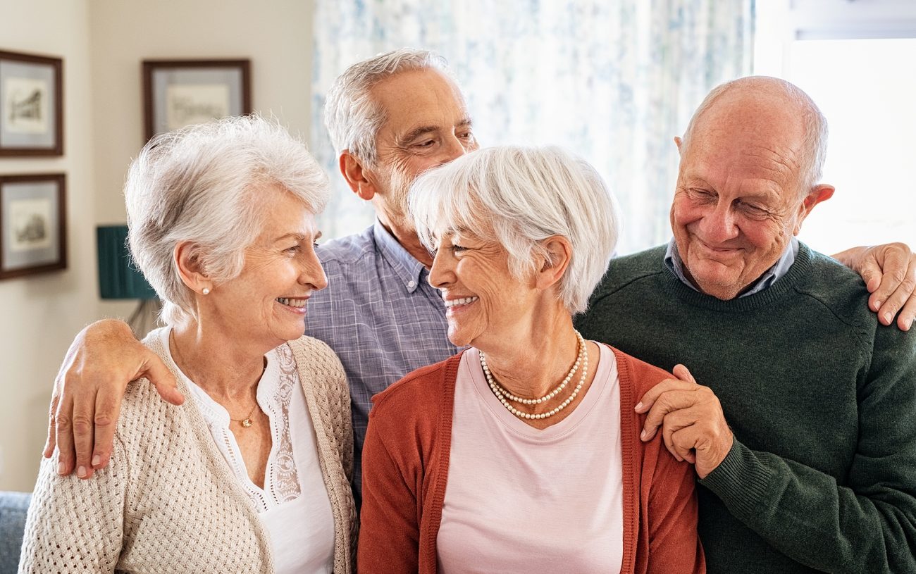 Happy senior couple with friends embracing and smiling at home. Cheerful senior men and beautiful women enjoying being together in eldercare facility centre: serene retirement and carefree concept.